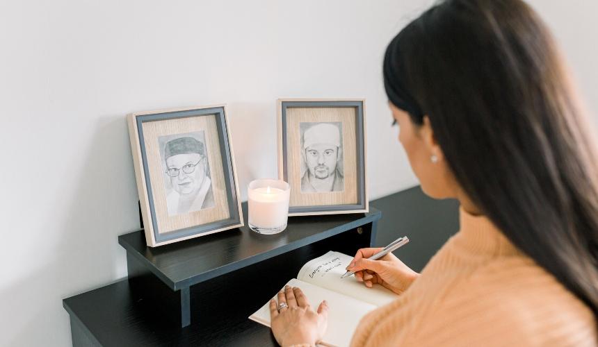 Dr Karan Juttla is shown from the back, writing in a notebook. In front of her are two framed pictures of a man and a woman with a candle sitting in front of the pictures.
