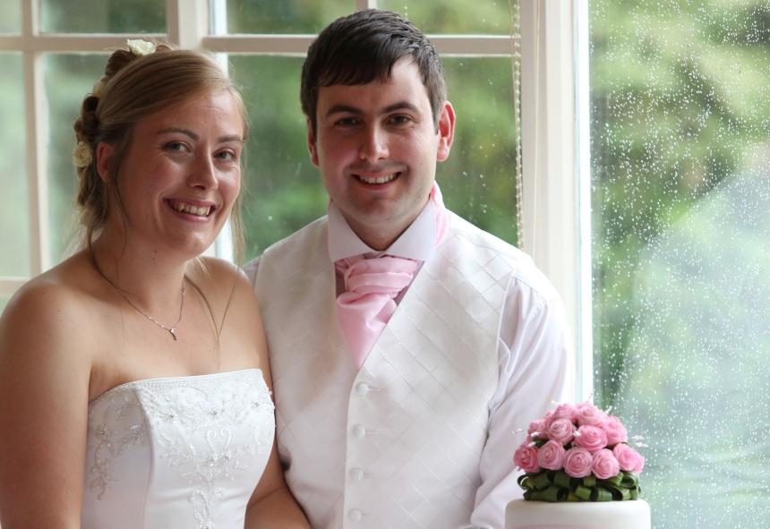 Caroline and Mark on their wedding day. Caroline is on the left in a white dress with lace embroidery. Mark is on the right in a white shirt and waistcoat. Both are smiling.
